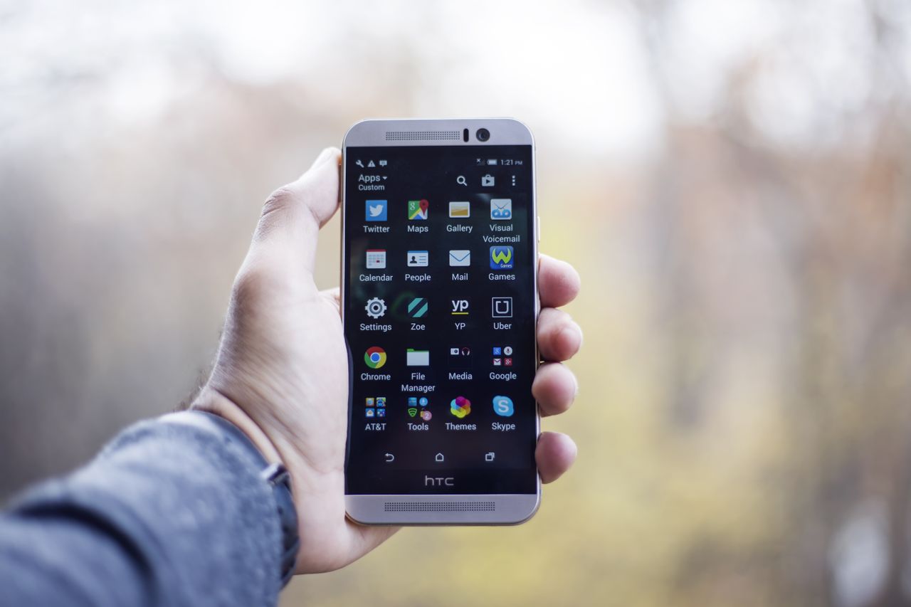 Services A close-up of a hand holding an HTC smartphone with visible apps on the screen, in an outdoor setting.